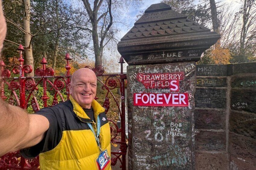 A man taking a selfie with a sign that reads 'STRAWBERRY FIELDS FOREVER' and 'BOB LAMARNE' on a post. 1