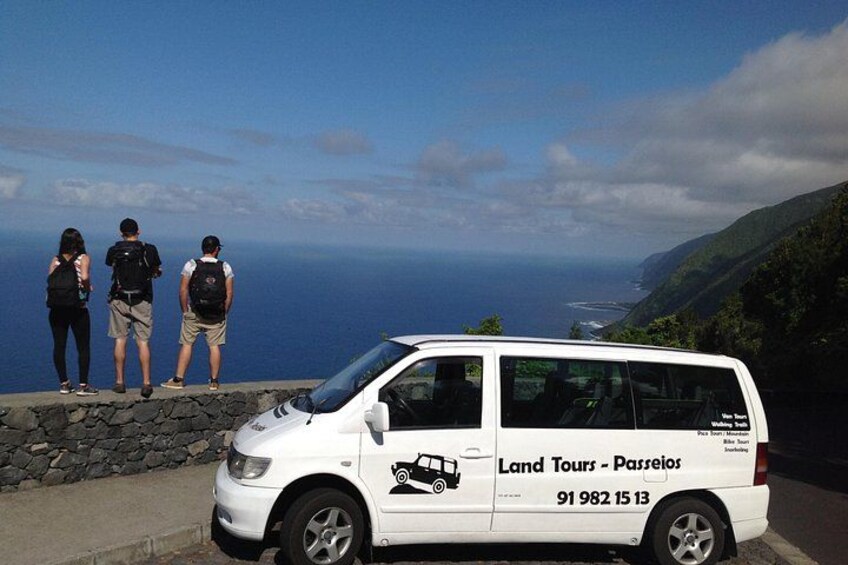 En hvid varevogn med teksten 'Land Tours - Passeios' og et telefonnummer holder parkeret ved vejen. 1