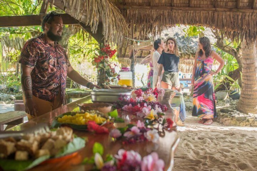 A tropical outdoor setting with a thatched-roof structure, a buffet table with food and flowers, and people socializing. 3