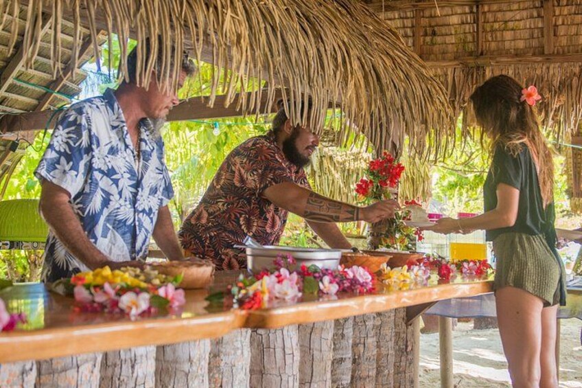 A tropical setting with a thatched roof structure, a wooden table adorned with flowers, and a person serving food. 2