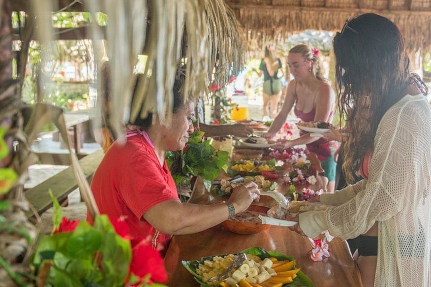 Un espace de restauration en plein air de style tropical, avec un toit de chaume, une table garnie de plats et des convives. 2