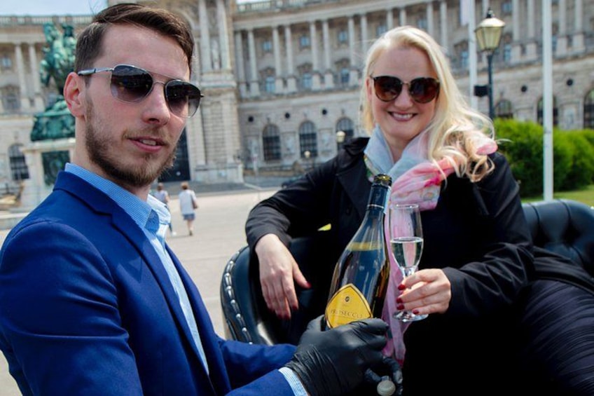Een man en een vrouw zitten buiten op een zwarte leren bank, met een fles champagne en een glas. 5