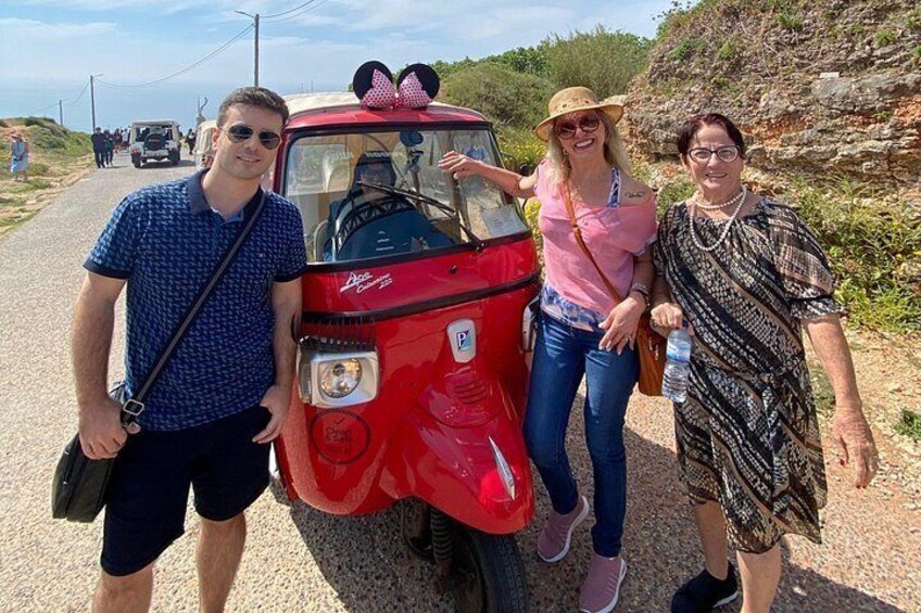 A red vintage car with a pink bow on the hood, a person in a blue shirt, and two other people standing beside it. 3