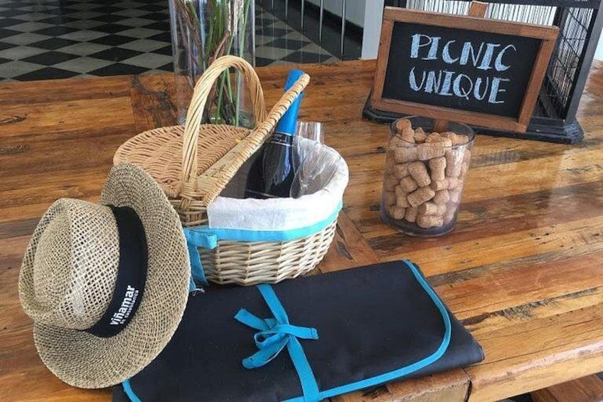 A picnic setup with a basket, a bottle, a hat, a blanket, and a jar of snacks on a wooden table. 1