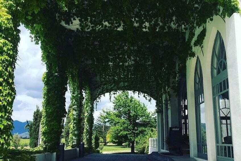 A covered walkway with arches, surrounded by greenery and a building with arched windows. 4
