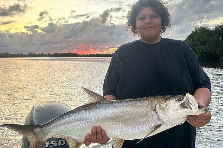 En person håller i en stor fisk vid en vattenyta, med en solnedgång i bakgrunden. 5