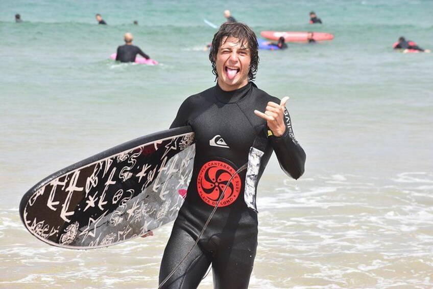 A person in a wetsuit holding a surfboard with graffiti, walking on a beach with other surfers in the background. 1