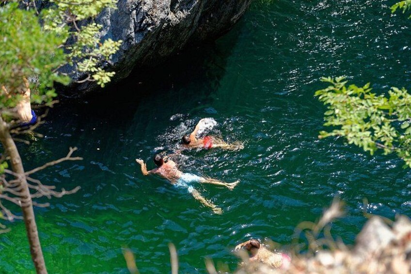 Des personnes nagent dans une étendue d’eau claire et verdoyante, entourée de rochers et de végétation. 3