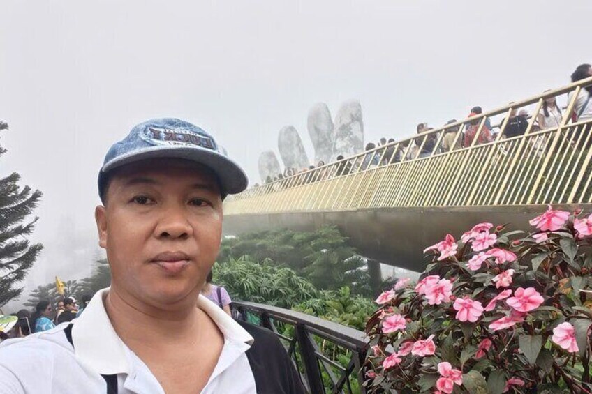 Eine Person steht vor einer Brücke, im Hintergrund leuchten rosa Blüten und Grünpflanzen. 2