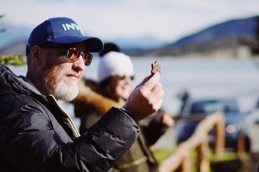 A person pointing with a finger, wearing a cap and jacket, with a body of water and hills in the background. 2