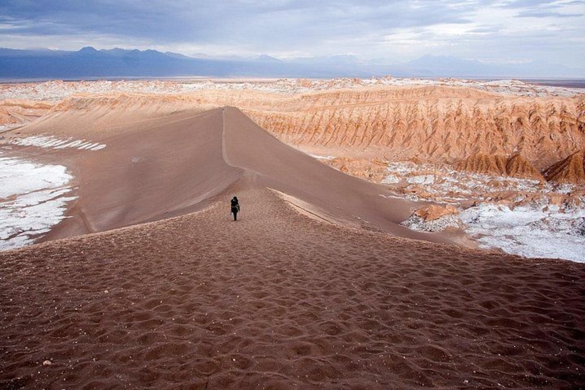 Una persona de pie al pie de una duna de arena enorme en un paisaje desértico, con montañas al fondo. 4