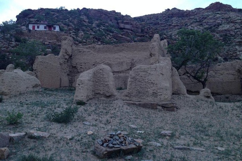 Ancient ruins with stone structures, a small building in the background, and a rocky hillside. 4