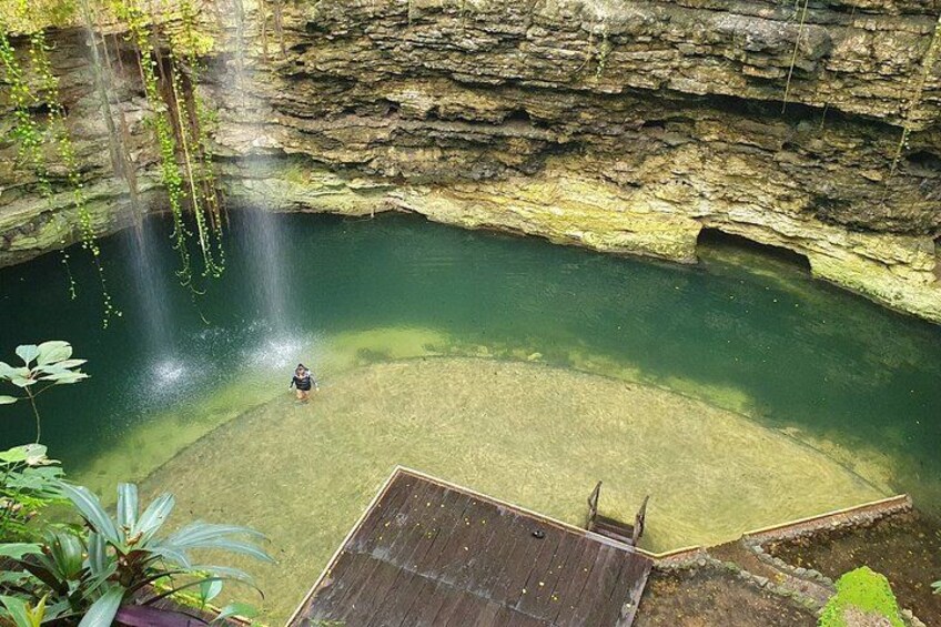 A person standing at the edge of a clear pool with a waterfall above, surrounded by rocky cliffs and greenery. 2