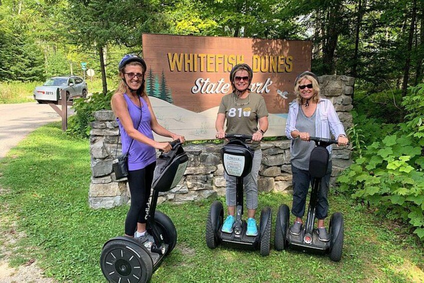 Three people on segways in front of a sign that reads 'WHITEFISH DUNES State Park'. 1