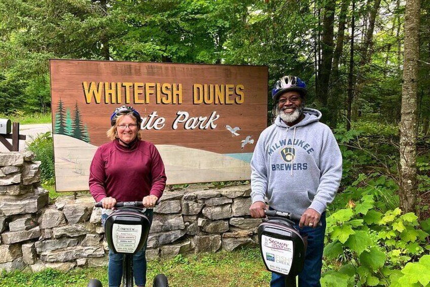 Deux personnes à vélo devant un panneau indiquant l’entrée du parc d’État de Whitefish Dunes. 3