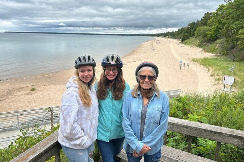 Trois personnes se tiennent sur une promenade en bois, surplombant une plage, avec une étendue d’eau en arrière-plan. 4