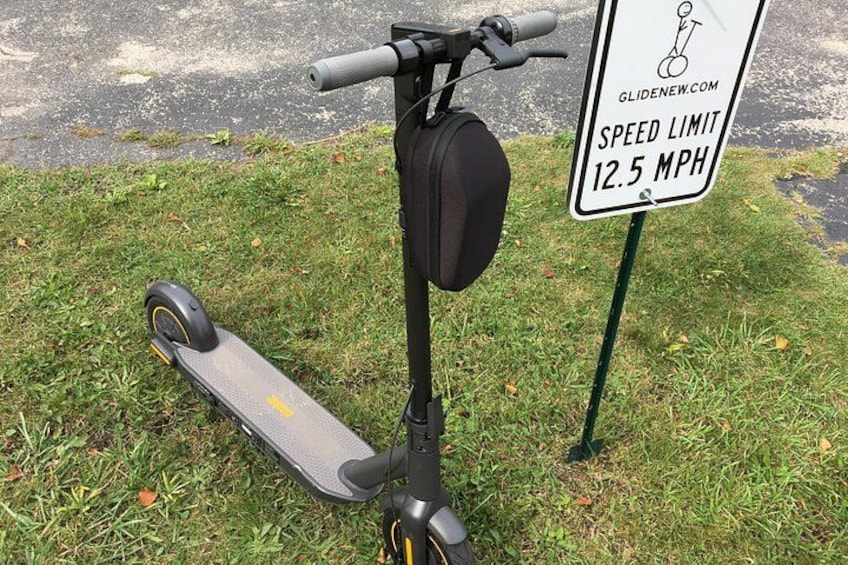 Una patineta eléctrica negra estacionada sobre césped junto a una señal de límite de velocidad que indica 20 kilómetros por hora. 2