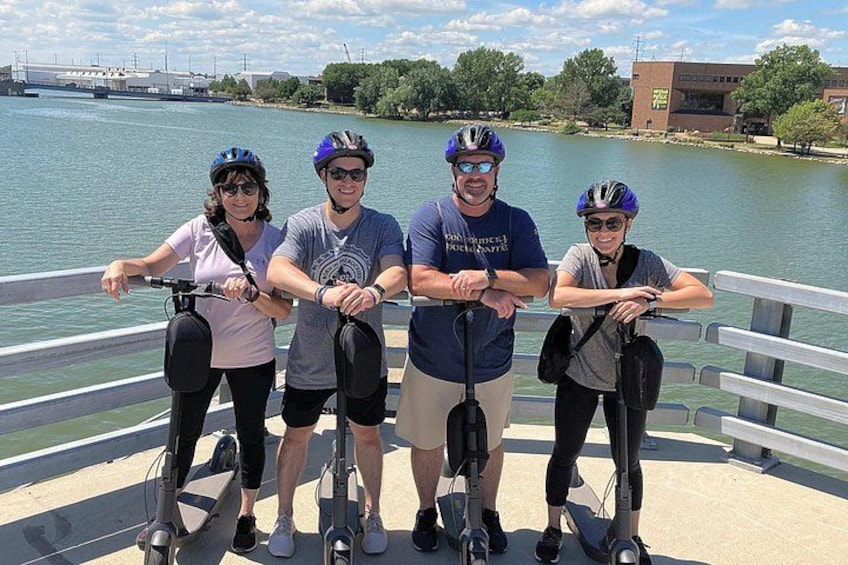 Cuatro personas en patinetes eléctricos junto a un río con un puente al fondo. 1
