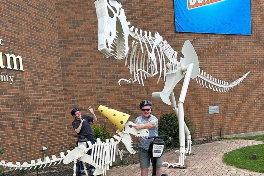 A large dinosaur skeleton sculpture, a brick building, and two people posing in front of it. 2