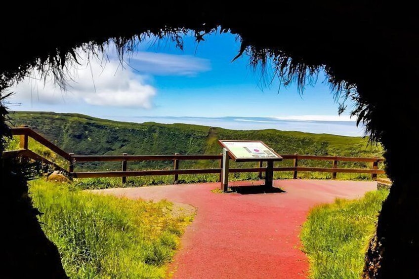 Un sendero rojo conduce a una pasarela de madera con un letrero, rodeado de colinas verdes y un cielo despejado. 3