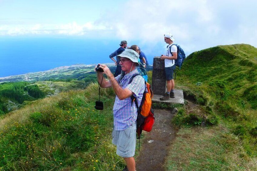 Un excursionista tomando una foto con una cámara, otro excursionista al fondo, y un sendero cubierto de hierba que conduce a un mirador con vista al mar. 5