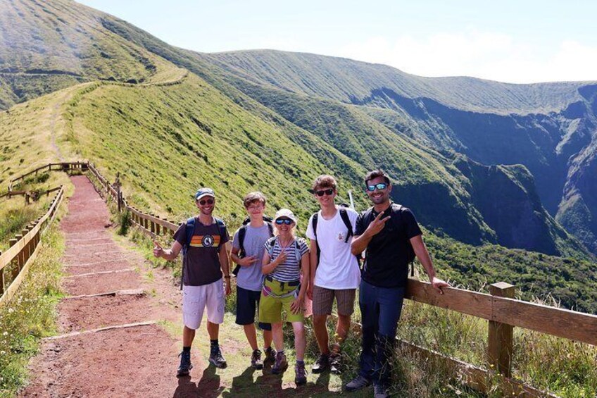 A group of people standing on a dirt path with a wooden railing, surrounded by green hills and a valley. 5