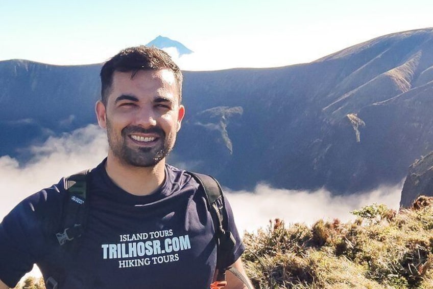 A person standing on a mountain with a backpack, wearing a t-shirt with 'Island Tours' and 'TRILHOSR.COM' text. 1