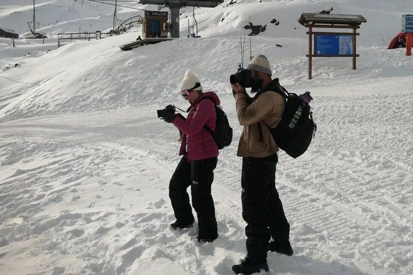 Zwei Personen machen Fotos in einer verschneiten Landschaft, im Hintergrund sind Seilbahnen und ein Wegweiser zu sehen. 2