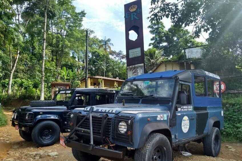 Un Land Rover Defender bleu est garé sur un chemin de terre, avec un SUV noir derrière lui. L’ensemble est entouré de verdure et se trouve à proximité d’un bâtiment sur lequel on peut voir une enseigne. 1