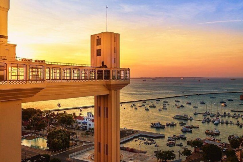 A coastal cityscape at sunset with a prominent bridge, a tower, and numerous boats in the harbor. 1