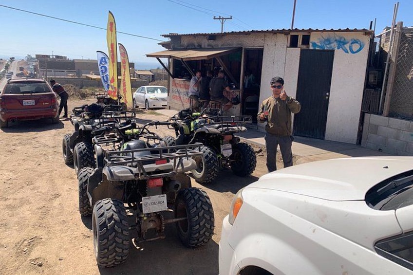ATVs parked in a dusty area with a white car in the foreground and a building with graffiti in the background. 4