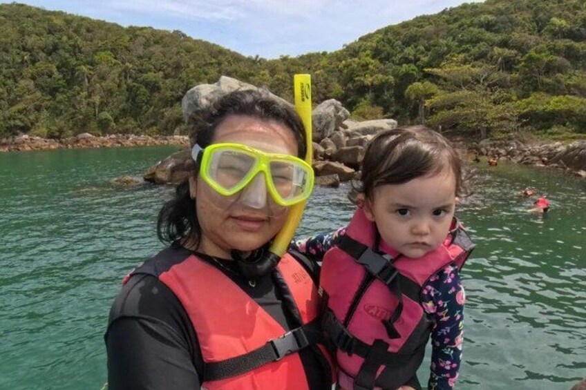 A person and a child wearing life jackets and snorkeling gear by a body of water with rocks and greenery in the background. 5