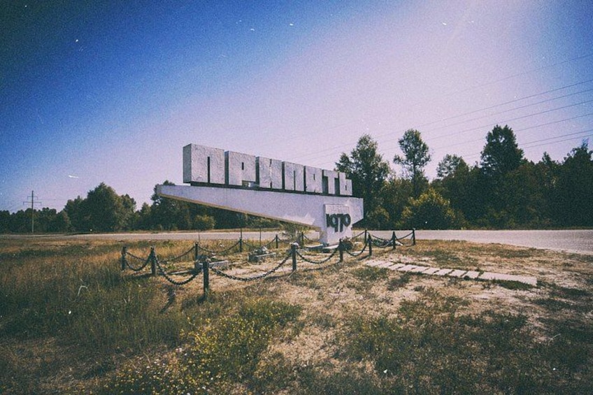 A white monument with inscriptions, surrounded by a chain-link fence, set in a grassy field with trees in the background. 1