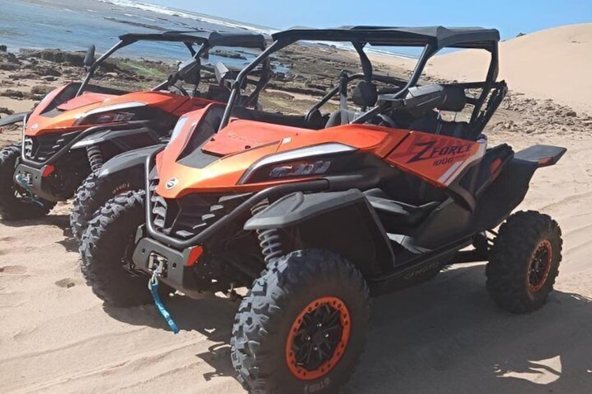 Drei orange-schwarz lackierte Geländewagen parken auf einem sandigen Strand, im Hintergrund eine felsige Küstenlinie. 2