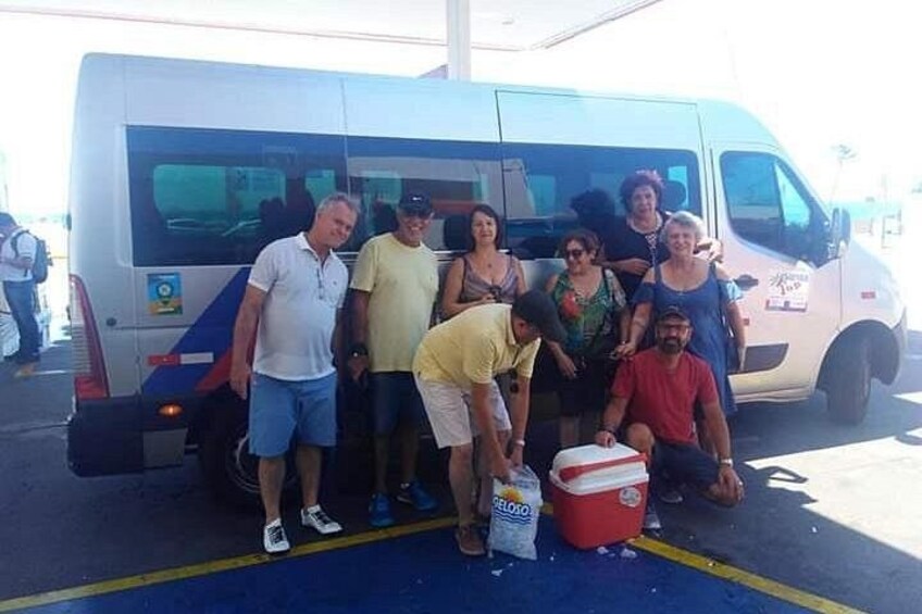 A group of people standing in front of a white van with a blue and red logo, a cooler, and a red container on the ground. 2