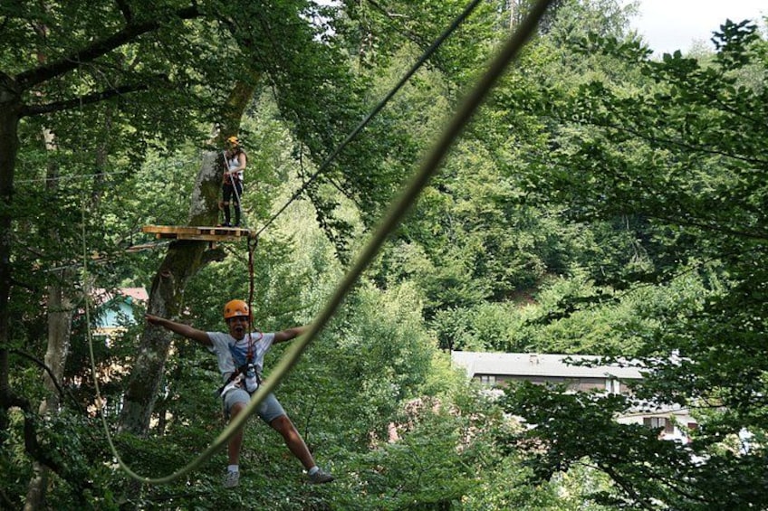 A person on a zip line, another on a platform, and a building in a forested area. 1
