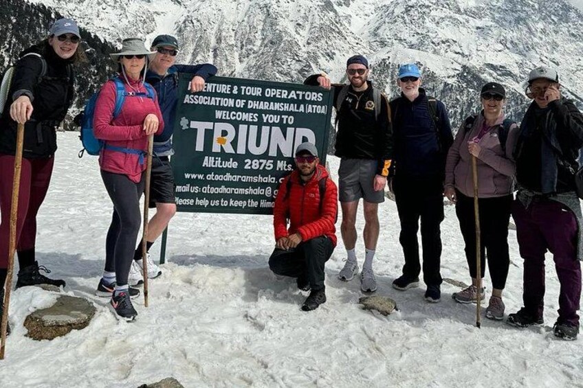 Een groep wandelaars staat in een besneeuwd landschap, bij een bord met de tekst 'TRIUNN' en de vermelding van een hoogte van 2875 meter. 4