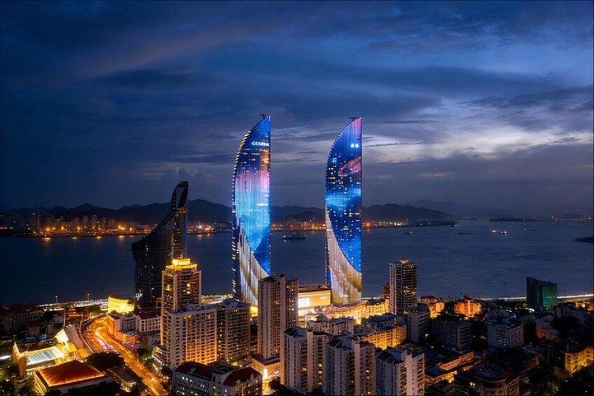Un paisaje urbano nocturno con dos rascacielos iluminados, una masa de agua y edificios circundantes. 3