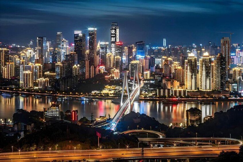 A city skyline at night with illuminated buildings, a prominent bridge, and a river reflecting the lights. 2