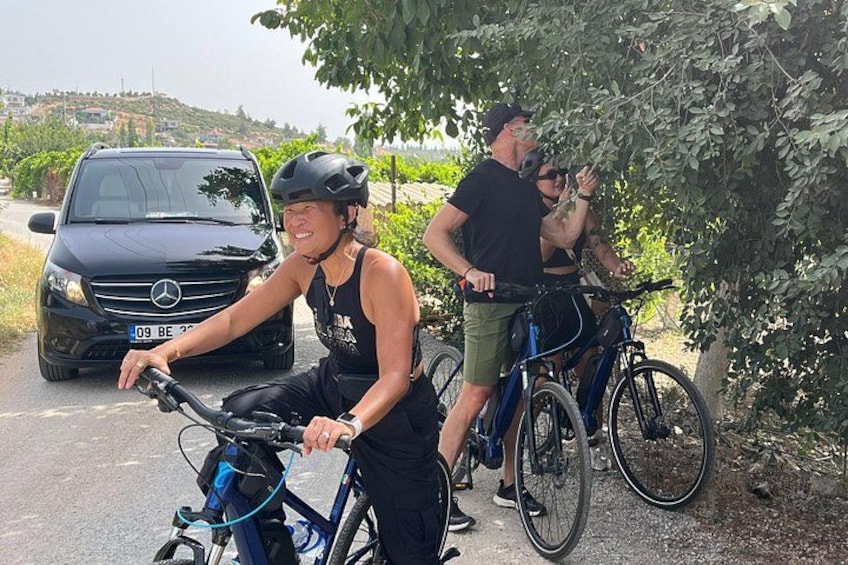 Eine Person fährt Fahrrad auf einem befestigten Weg, ein geparktes Auto und eine weitere Person steht neben einem Baum. 2