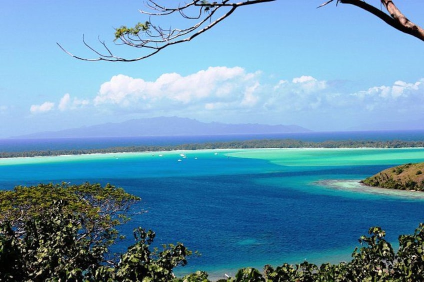 A coastal landscape with clear turquoise waters, a distant mountain range, and dense vegetation in the foreground. 2