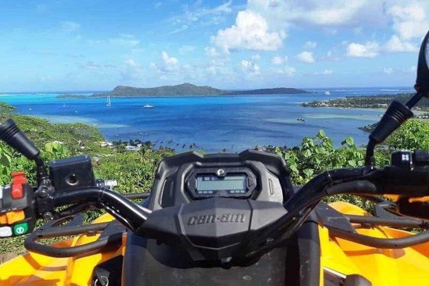 En Can-Am ATV med digital skærm, omgivet af frodig vegetation og udsigt over et kystlandskab med vand og fjerne øer. 1
