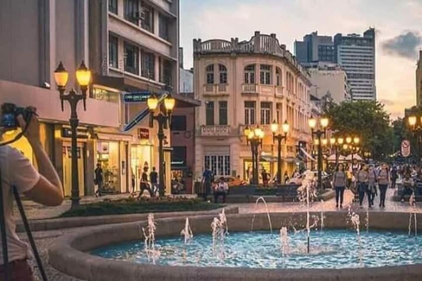 A public square with a fountain, buildings, and street lamps. 2