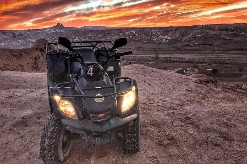 A quad bike with headlights on, parked on a dirt surface with a sunset in the background. 3