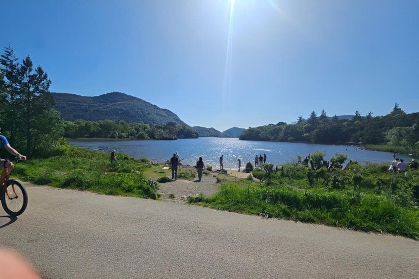 A lake with people walking and cycling along the shore, surrounded by greenery and hills. 4