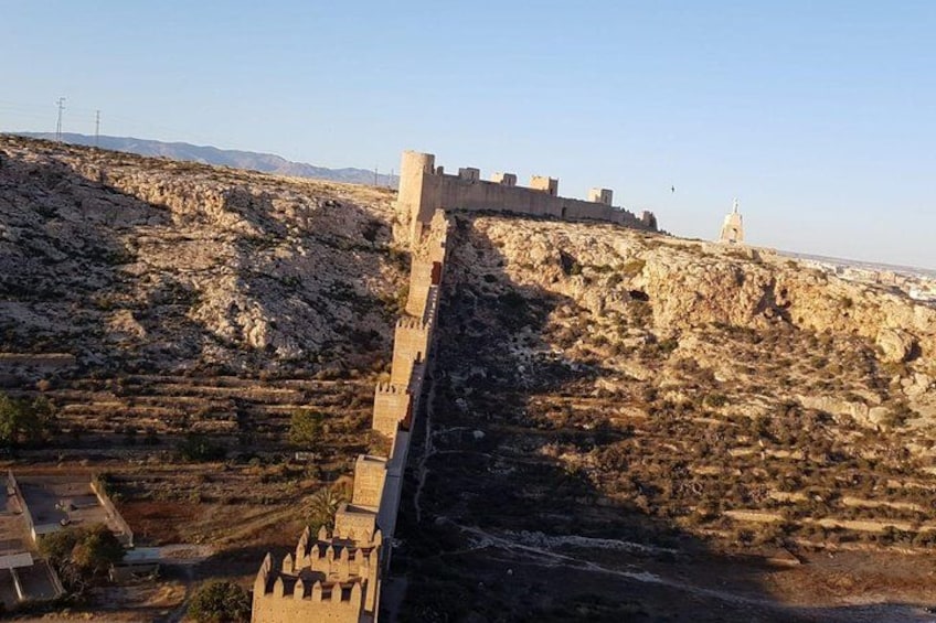 A historical stone wall with a tower, a rocky hillside, and a distant bell tower. 5