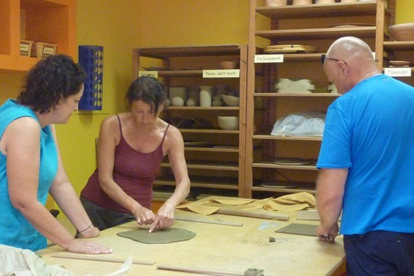 Three people engaged in pottery making, with clay wheels and tools on a table, and shelves with pottery in the background. 5