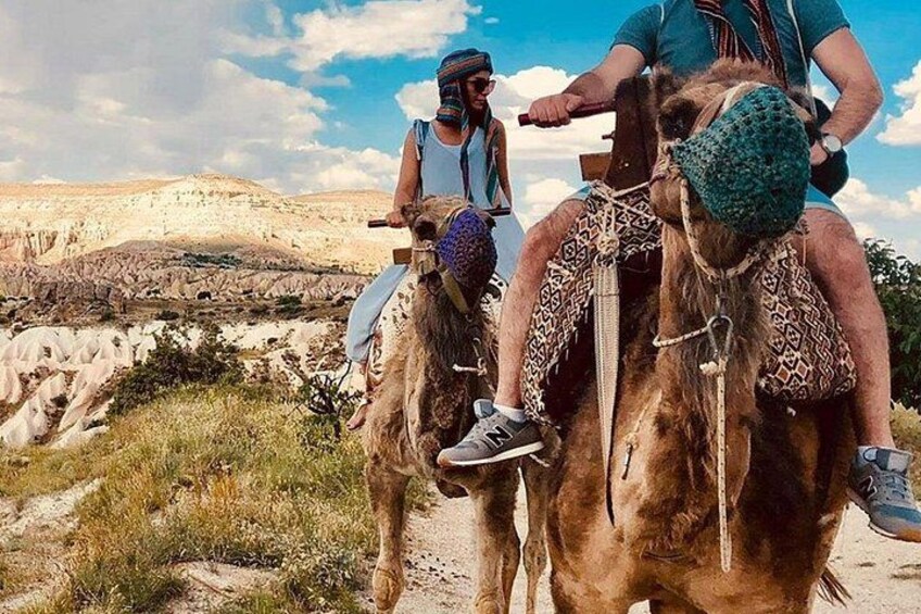 Two people riding camels on a dirt path with a rocky landscape in the background. 2