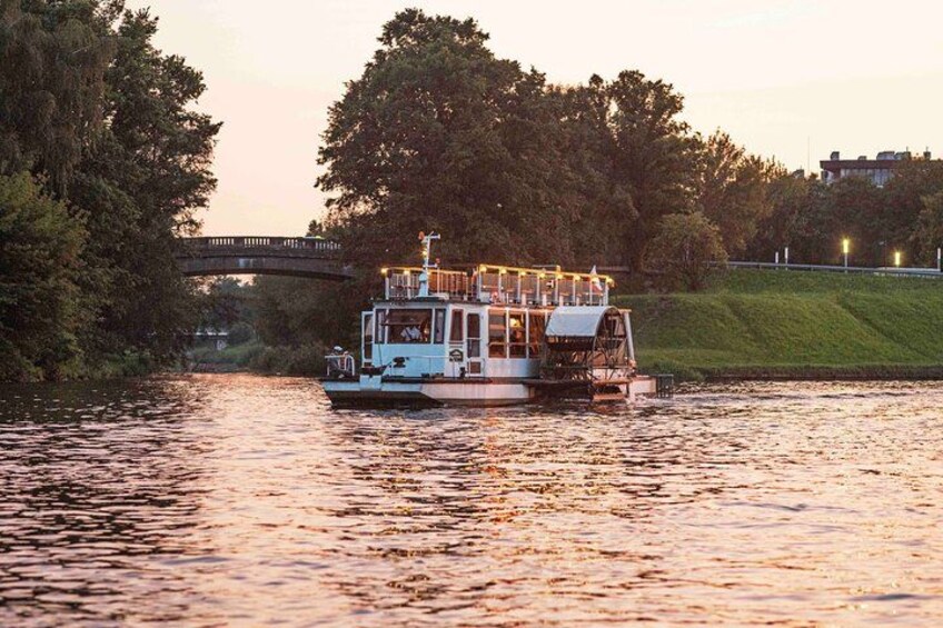 A riverboat with a covered deck and lights, moving under a bridge with trees and a grassy bank in the background. 3