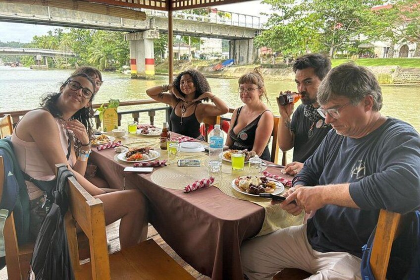 A group of people dining at a table by a river with a bridge in the background. 4